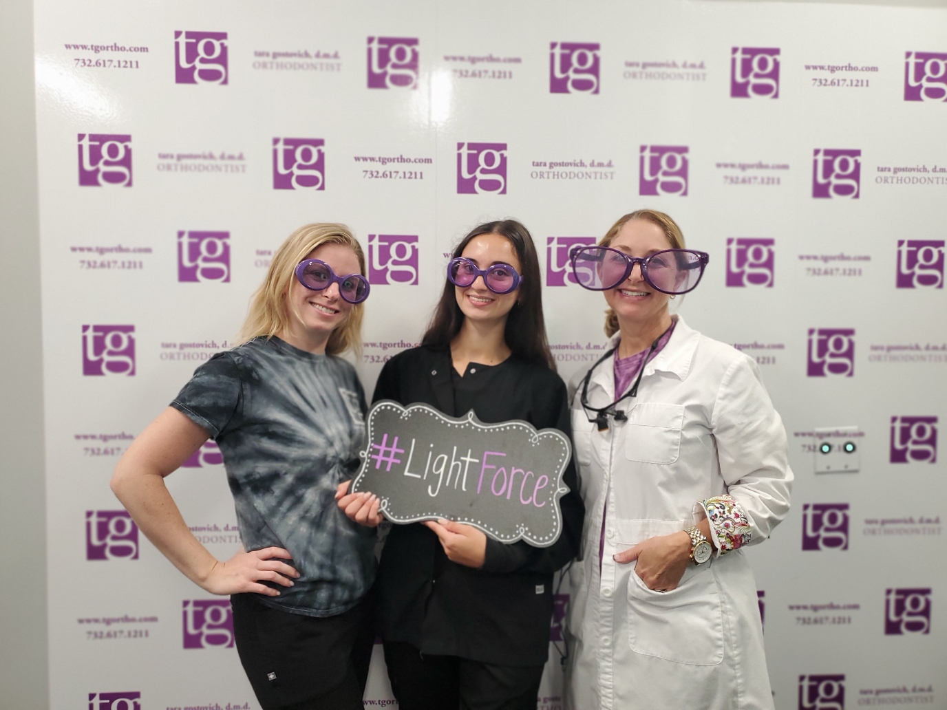 Three women wearing large purple sunglasses, smiling and holding up a LightForce sign.