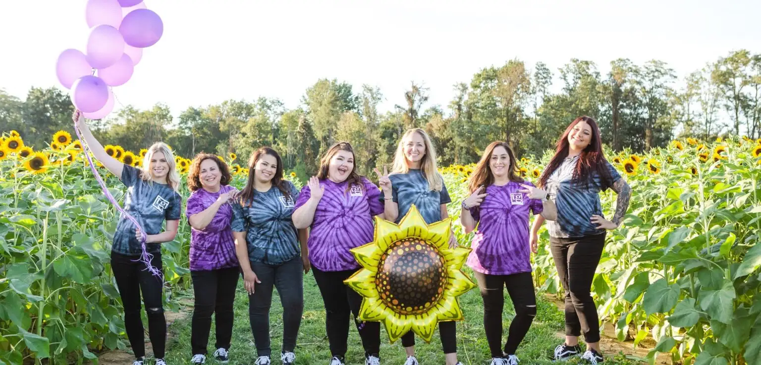 Seven individuals in tie-dyed T-shirts standing together in a vibrant sunflower field.